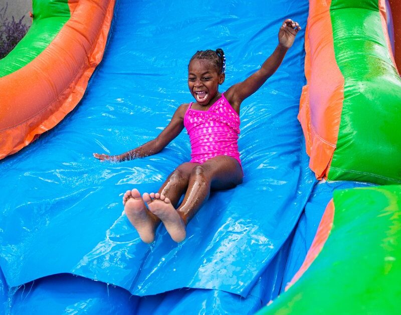 A young child wearing a pink bathing suit sliding down a large inflatable bounce house water slide outside.