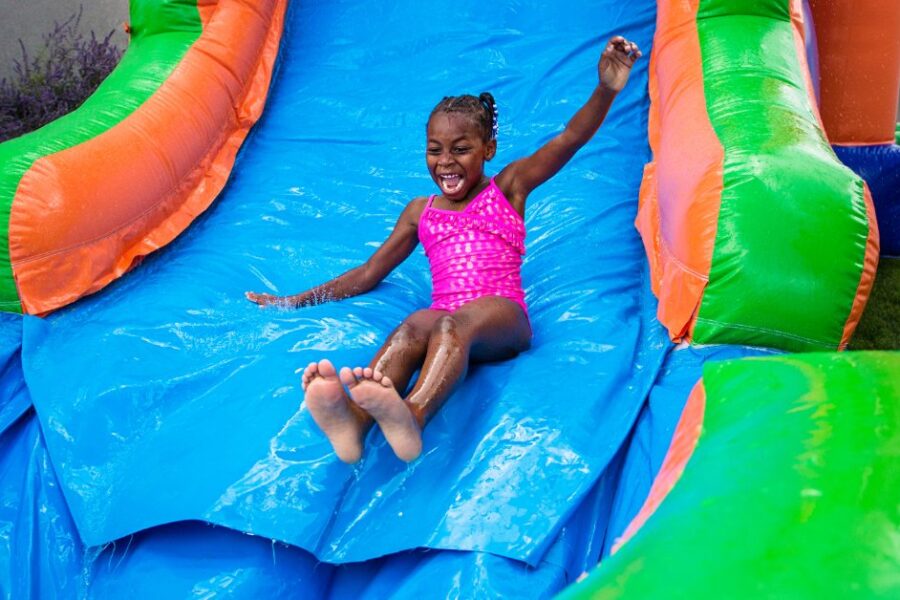 A young child wearing a pink bathing suit sliding down a large inflatable bounce house water slide outside.