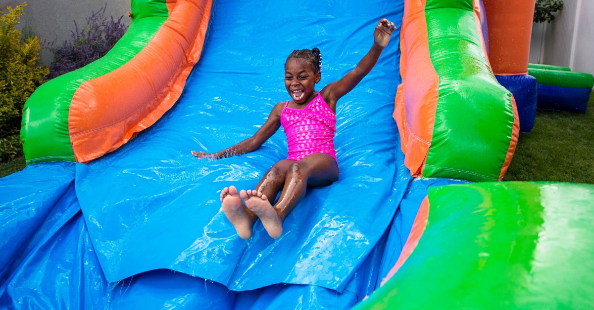 A young child wearing a pink bathing suit sliding down a large inflatable bounce house water slide outside.