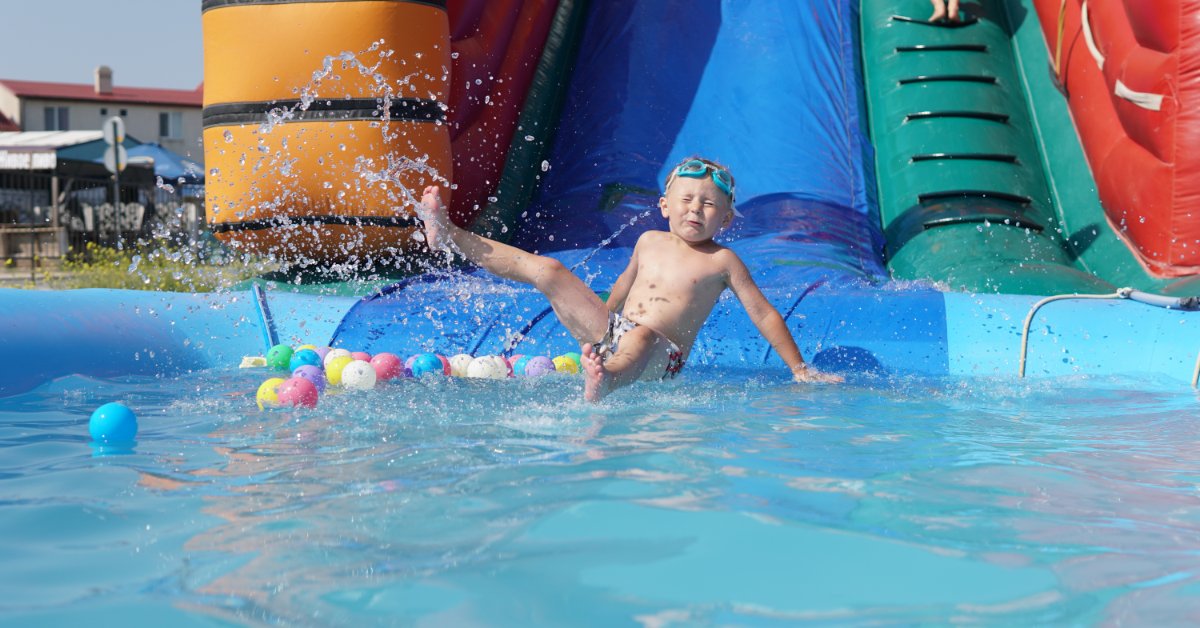 A young child wearing blue goggles sliding down a blue inflatable water slide leading into a small pool.