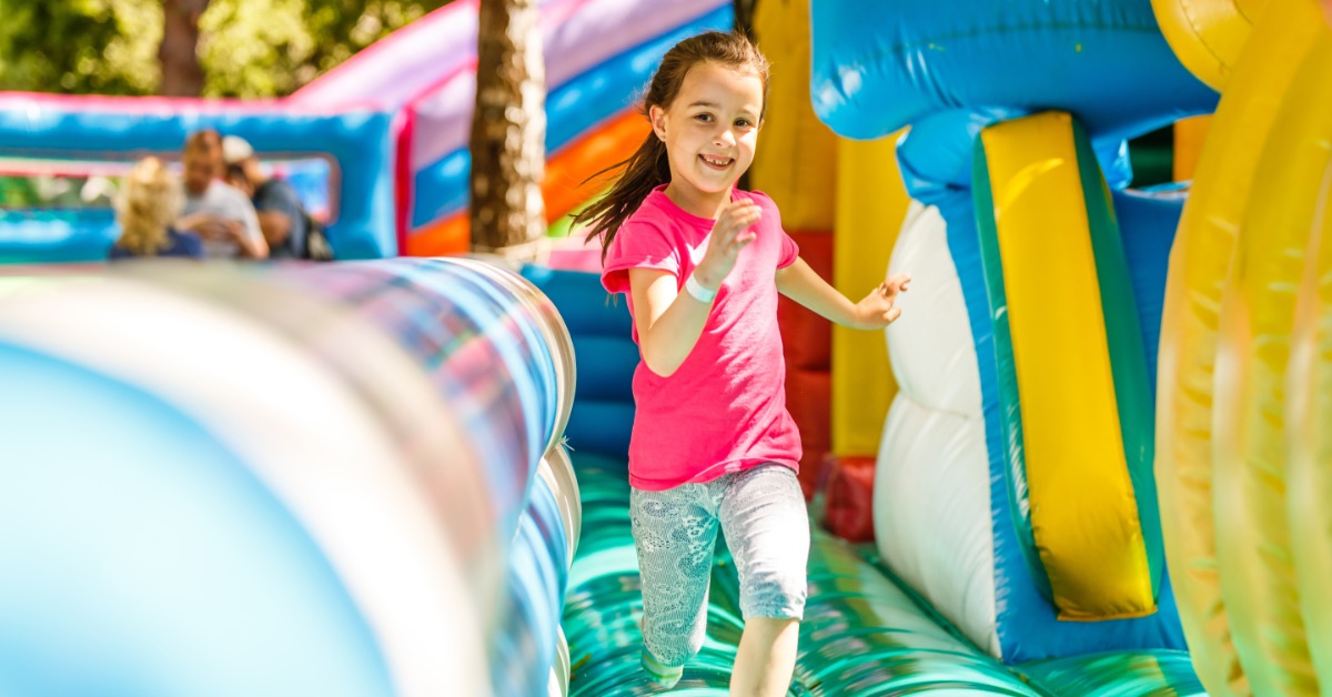 A child wearing a bright pink shirt smiling and running on a colorful inflatable obstacle course outside.