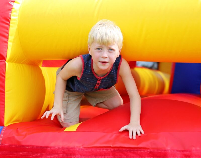 A child wearing a blue-and-red shirt crawls out of a large inflatable obstacle course outside on a sunny day.