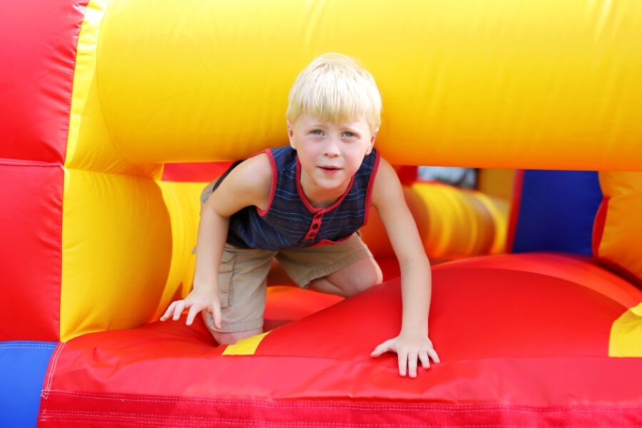 A child wearing a blue-and-red shirt crawls out of a large inflatable obstacle course outside on a sunny day.