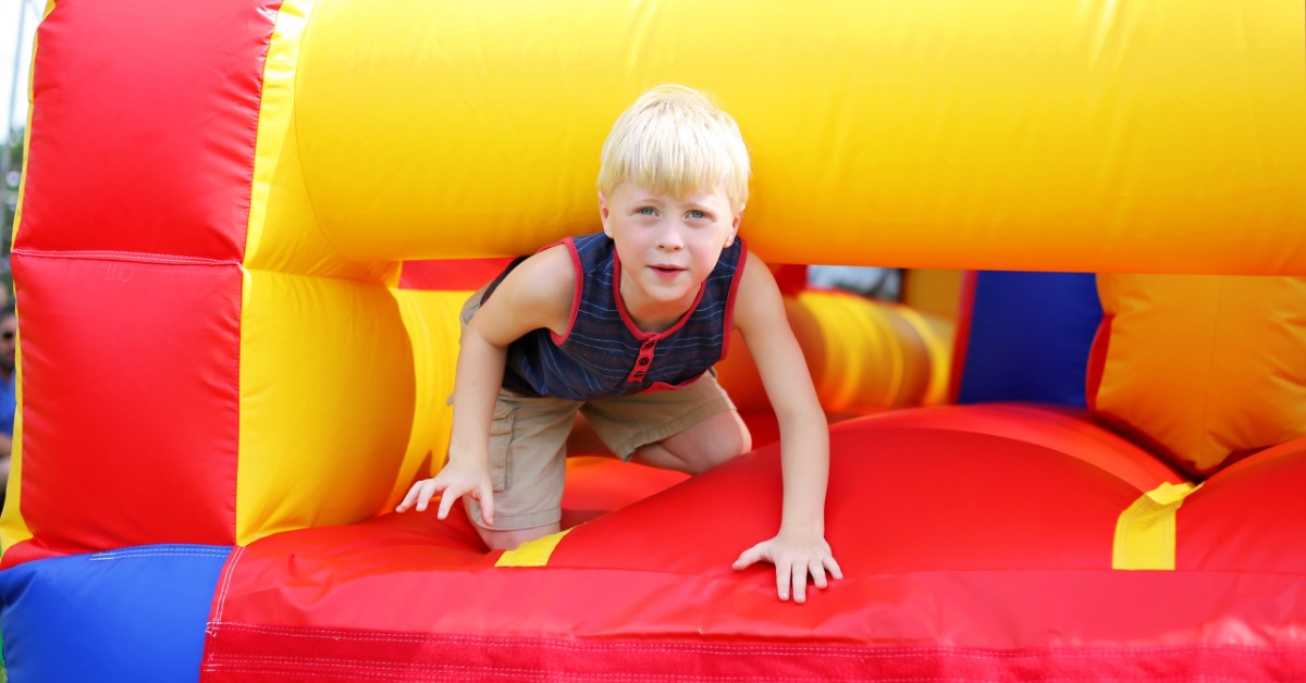 A child wearing a blue-and-red shirt crawls out of a large inflatable obstacle course outside on a sunny day.