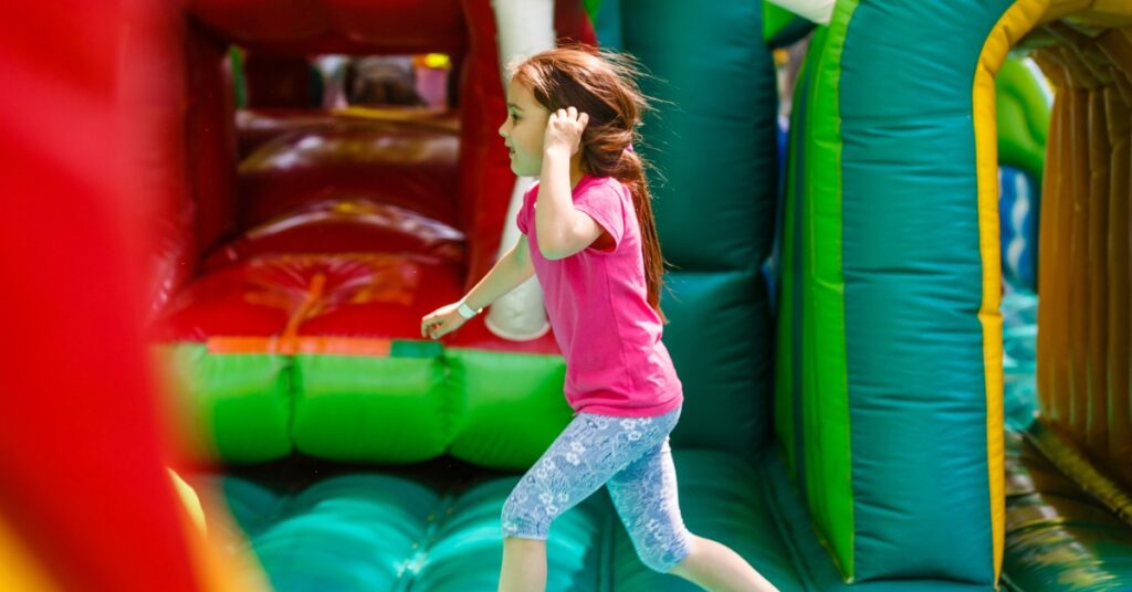 A child wearing a pink shirt runs next to two tunnels on a large inflatable obstacle course outside.