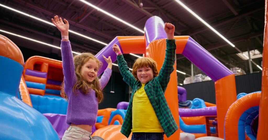 Two kids raise their arms and smile in a massive, colorful bounce house. The bounce house is in an indoor space.