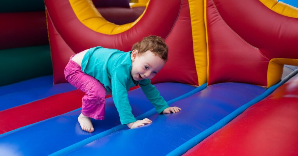 A toddler wearing a bright blue shirt and pink pants excitedly climbs on a large, colorful bounce house outside.