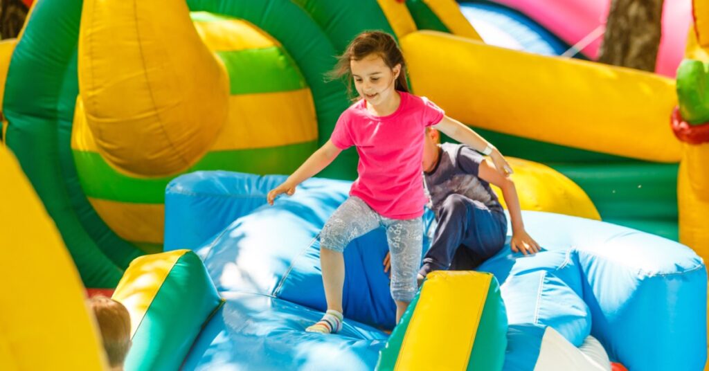 Two children climbing over a short blue obstacle in an outdoor inflatable obstacle course on a sunny day.