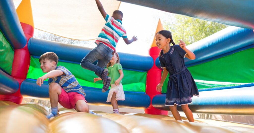 Four children jumping in a large roofless inflatable bounce house near a tall tree outside on a sunny day.