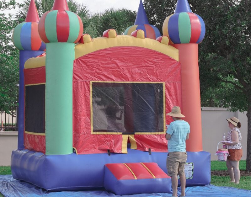 Two people standing outside a colorful bounce house on a blue tarp at a party in a yard during the daytime.