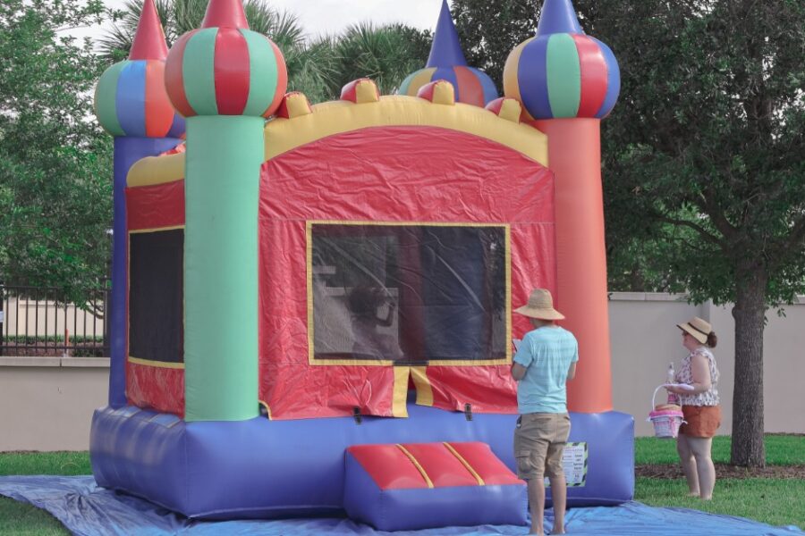Two people standing outside a colorful bounce house on a blue tarp at a party in a yard during the daytime.