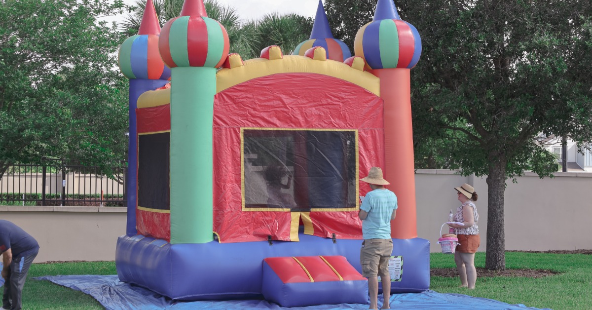 Two people standing outside a colorful bounce house on a blue tarp at a party in a yard during the daytime.